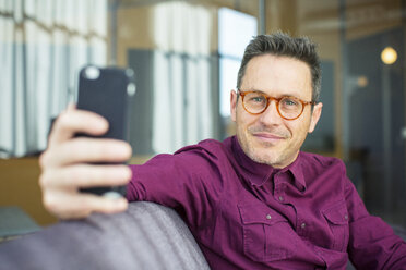 Porträt eines Geschäftsmannes, der ein Mobiltelefon benutzt, während er auf dem Sofa im Büro sitzt - CAVF47241