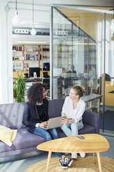 Female colleagues with laptop computer discussing on sofa in office - CAVF47245