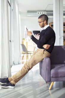 Seitenansicht eines Geschäftsmannes, der sein Smartphone benutzt, während er sich auf dem Sofa im Büro anlehnt - CAVF47250