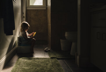 Seitenansicht eines kleinen Mädchens auf dem Töpfchen, während es ein Eis am Stiel in der Dunkelkammer zu Hause hält - CAVF47309