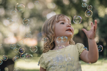 Cute baby girl reaching for bubbles flying in air at park - CAVF47315