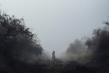 Girl standing on field against clear sky while being wrapped in blanket during foggy weather - CAVF47319