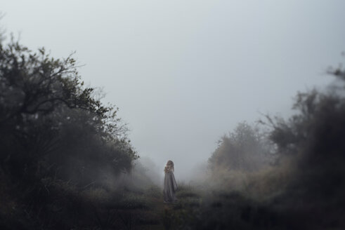 Mädchen steht auf einem Feld gegen den klaren Himmel und ist in eine Decke eingewickelt, während es neblig ist - CAVF47319