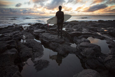 Rückansicht eines Surfers, der ein Surfbrett trägt, während er bei Sonnenuntergang am felsigen Ufer steht - CAVF47336