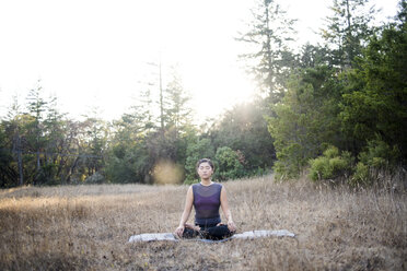 Woman meditating while sitting on exercise mat against clear sky at field - CAVF47376