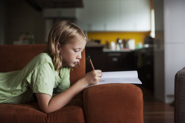 Seitenansicht eines Jungen, der in ein Buch schreibt, während er zu Hause auf dem Sofa liegt - CAVF47457