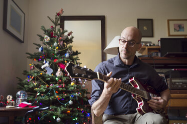 Mann spielt Gitarre, während er zu Hause am Weihnachtsbaum sitzt - CAVF47492