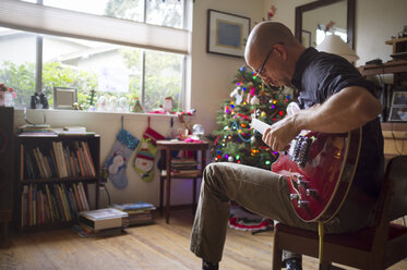 Mann spielt Gitarre und sitzt zu Hause auf einem Stuhl während der Weihnachtszeit - CAVF47494