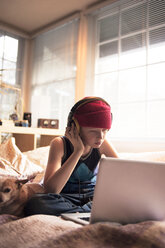 Junge hört Musik mit Kopfhörern, während er einen Laptop auf dem Bett benutzt - CAVF47505
