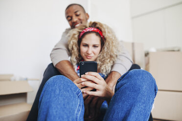 Smiling couple using smart phone while sitting at new home - CAVF47552