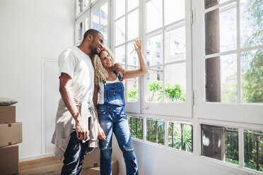 Smiling couple looking at keys while standing by window at home - CAVF47563