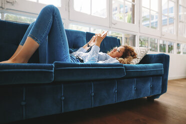 Junge Frau in voller Länge, die ihr Smartphone benutzt, während sie zu Hause auf dem Sofa liegt - CAVF47592