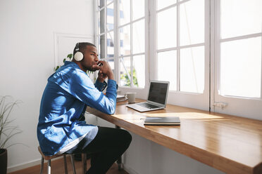 Side view of thoughtful man listening music while sitting with laptop computer at wooden table by window - CAVF47595