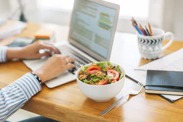 Cropped image of businesswoman blogging through laptop computer by salad bowl on wooden table at home office - CAVF47598