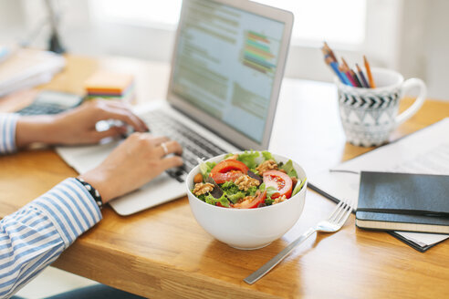 Cropped image of businesswoman blogging through laptop computer by salad bowl on wooden table at home office - CAVF47598