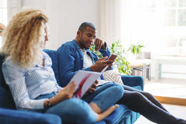 Frau liest eine Zeitschrift, während der Mann auf dem Sofa zu Hause sein Smartphone benutzt - CAVF47609