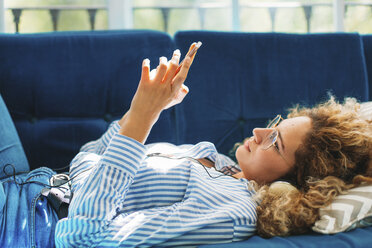 Seitenansicht einer Frau, die ein Smartphone benutzt, während sie zu Hause auf dem Sofa liegt - CAVF47612