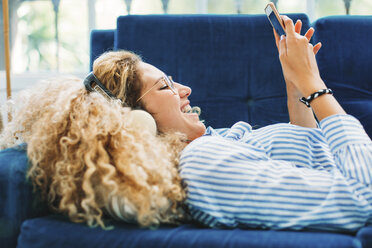 Seitenansicht einer fröhlichen Frau, die ein Smartphone benutzt, während sie zu Hause auf dem Sofa liegt - CAVF47613