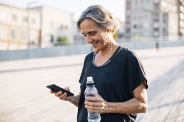 Glückliche Frau schaut auf ihr Smartphone und hält eine Wasserflasche - CAVF47632