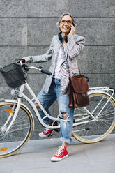 Happy woman talking on smart phone with bicycle against wall on footpath - CAVF47642