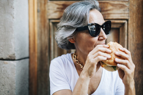 Frau mit Sonnenbrille isst Burger und sitzt gegen die Tür - CAVF47655