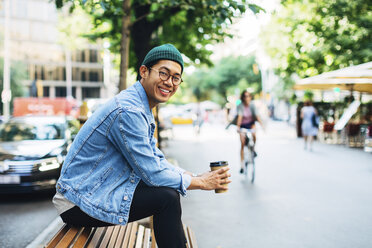 Porträt eines glücklichen Mannes, der einen Einwegbecher hält, während er auf einer Bank am Straßenrand sitzt - CAVF47693