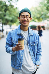Portrait of happy man with hand in pocket holding disposable cup at sidewalk - CAVF47694