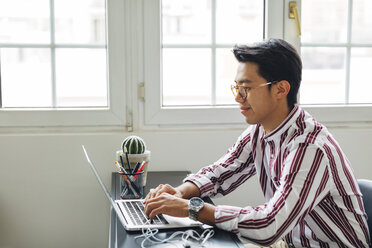 Geschäftsmann, der an einem Laptop arbeitet, während er im Büro am Fenster sitzt - CAVF47721