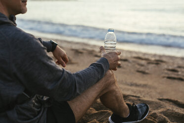 Low section of man holding water bottle while sitting at beach - CAVF47735