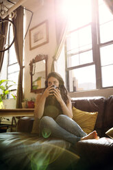 Frau in voller Länge beim Teetrinken auf dem Sofa zu Hause an einem sonnigen Tag - CAVF47761