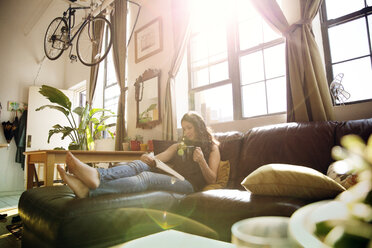 Woman reading book while having tea on sofa at home during sunny day - CAVF47762