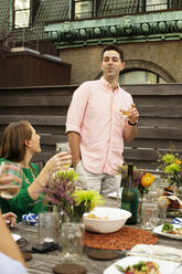 Woman looking at man holding drink while enjoying at table on building terrace - CAVF47944