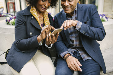 Smiling couple using smart phone while sitting on seat in city - CAVF48129