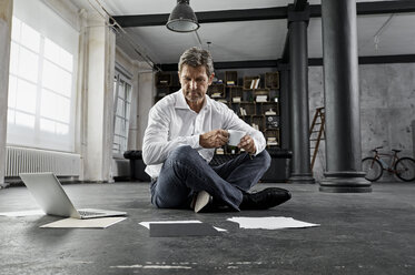 Mature man sitting on ground with laptop and notes in loft - PDF01582