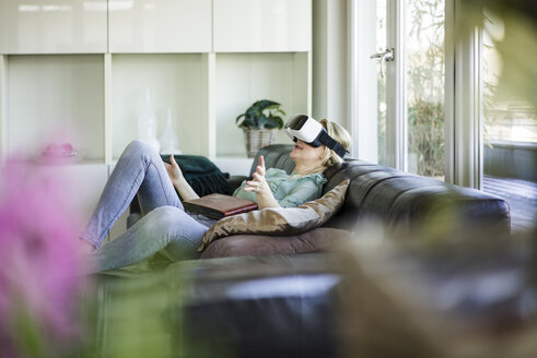 Frau auf der Couch liegend mit Fotoalbum und Virtual-Reality-Brille - MOEF01059