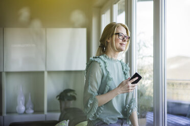 Portrait of content woman with smartphone looking out of window - MOEF01063