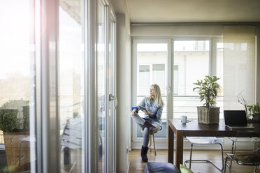 Woman with tablet sitting on stool looking out of window - MOEF01068