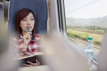 Thoughtful young woman writing in journal and looking out window on passenger train - CAIF20222