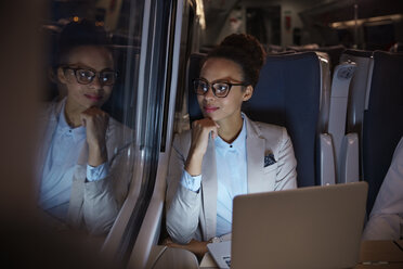 Selbstbewusste, nachdenkliche Geschäftsfrau, die nachts im Personenzug aus dem Fenster schaut und am Laptop arbeitet - CAIF20250