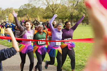 Enthusiastic female runners in tutus crossing charity run finish line in park, celebrating - CAIF20309