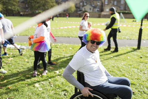 Porträt eines lächelnden, verspielten Mannes im Rollstuhl mit Clownsperücke bei einem Wohltätigkeitsrennen in einem sonnigen Park, lizenzfreies Stockfoto