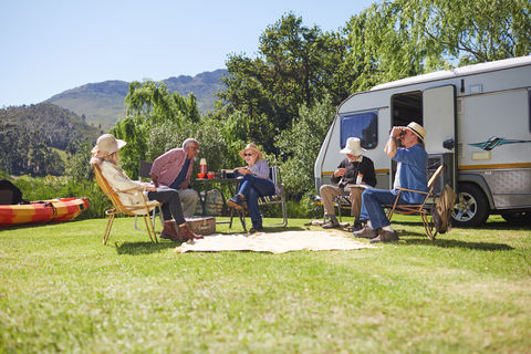 Aktive ältere Freunde entspannen sich vor einem Wohnmobil auf einem sonnigen Campingplatz, lizenzfreies Stockfoto