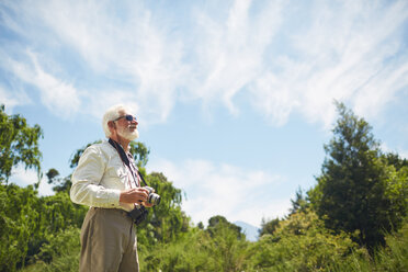 Neugieriger aktiver älterer Mann mit Digitalkamera, der zu den sonnigen Bäumen und dem Himmel aufschaut - CAIF20388