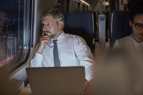 Ernster, nachdenklicher Geschäftsmann, der an seinem Laptop arbeitet und aus dem Fenster eines nächtlichen Personenzugs schaut - CAIF20407