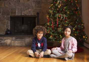 Porträt von Geschwistern, die zu Hause vor dem Weihnachtsbaum sitzen - CAVF48277