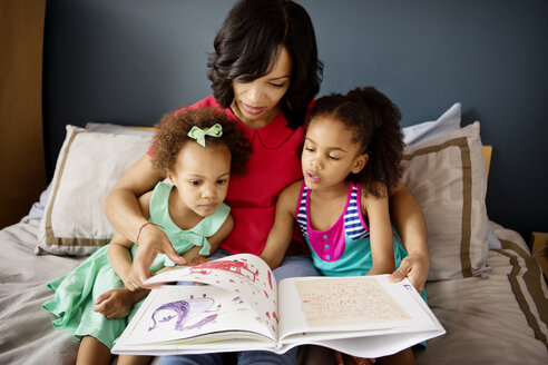 Mutter und Kinder schauen in ein Buch, während sie zu Hause auf dem Bett sitzen - CAVF48346