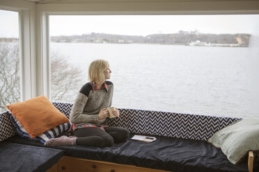 Thoughtful woman having coffee while looking at lake through window during winter - CAVF48353