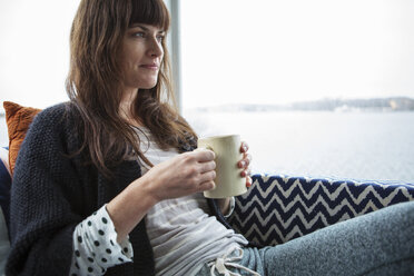 Thoughtful smiling woman having coffee while sitting by glass window at home - CAVF48354