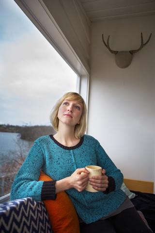 Nachdenkliche Frau, die einen Kaffee trinkt und durch ein Fenster in ihrem Haus schaut, lizenzfreies Stockfoto