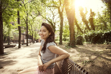 Lächelnde Frau sitzt auf Bank gegen Bäume während sonnigen Tag im Park - CAVF48392